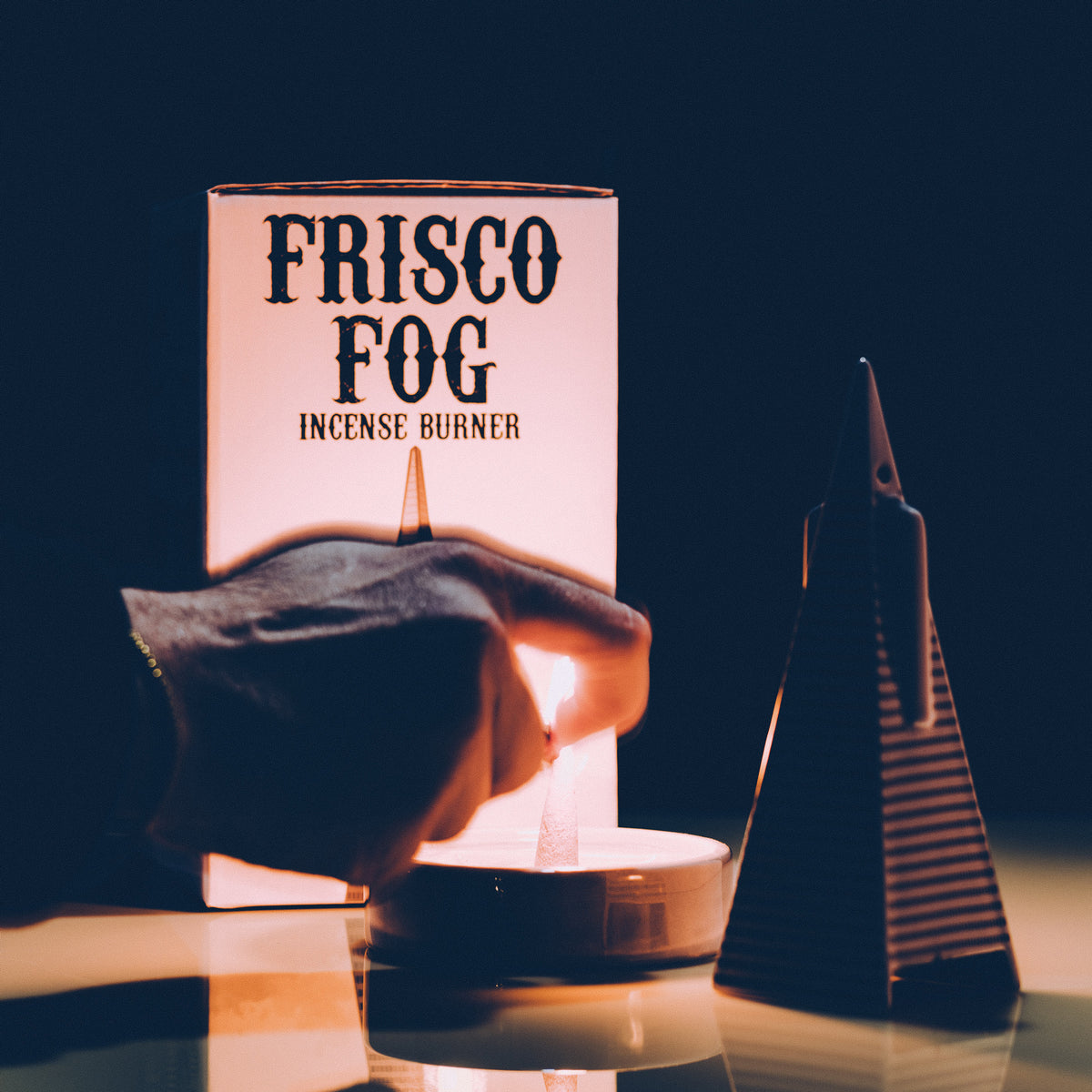 A hand holds a lit incense burner in front of a glowing box labeled 'FRISCO FOG INCENSE BURNER,' with a dark triangular incense holder beside it on a reflective surface.