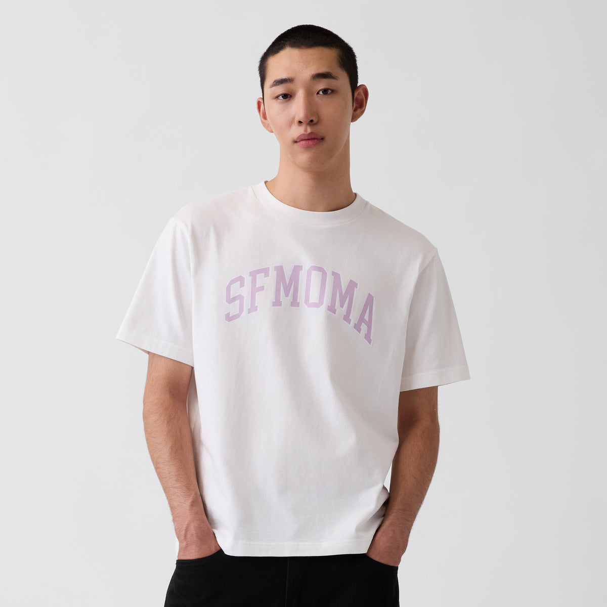 An off-white short-sleeve crewneck t-shirt featuring the word 'SFMOMA' printed in an arched, dusty pink collegiate-style font across the chest, displayed on a clean white background.