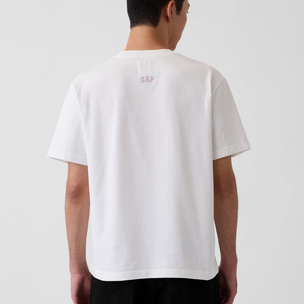 A man viewed from the back wearing a white short-sleeve t-shirt with a small pink 'GAP' logo centered on the upper back, paired with black pants against a light gray studio background.