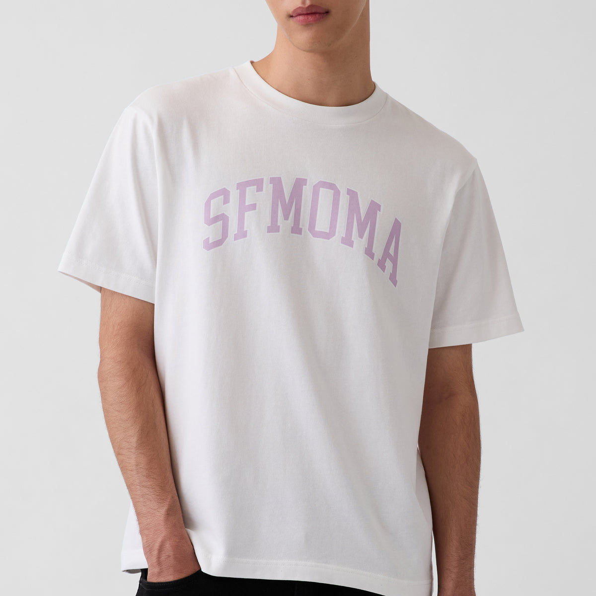 A white crewneck t-shirt with 'SFMOMA' printed in arched lavender-purple varsity letters across the chest, worn by a model against a light gray background.