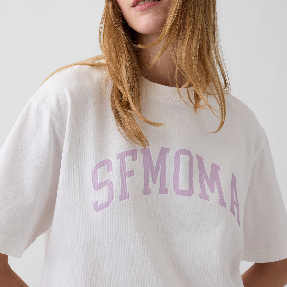 A close-up of a woman wearing a white oversized T-shirt with the word 'SFMOMA' printed across the chest in a light lavender-pink arched font.