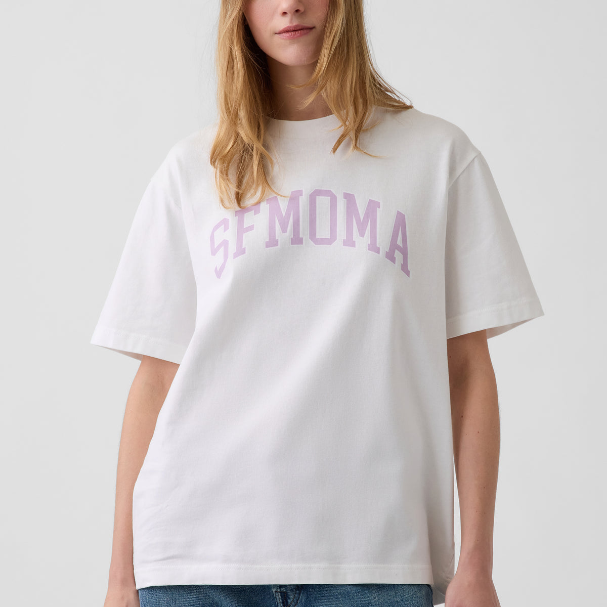 A woman wearing an oversized white t-shirt with 'SFMOMA' printed in a light purple collegiate-style font across the chest, paired with blue jeans against a light gray background.