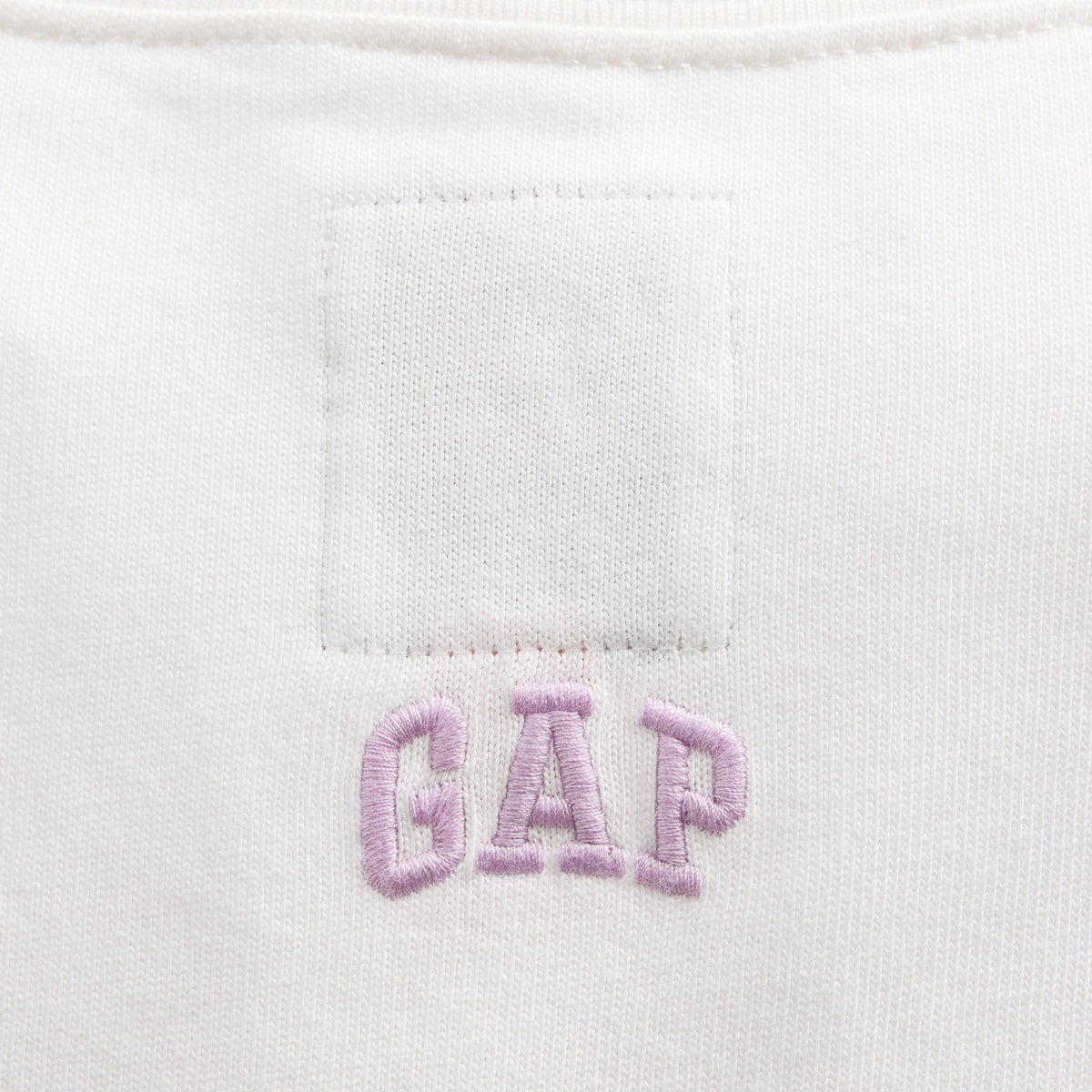 Close-up of a white garment fabric with a rectangular stitched patch and light pink embroidered 'GAP' lettering below it.
