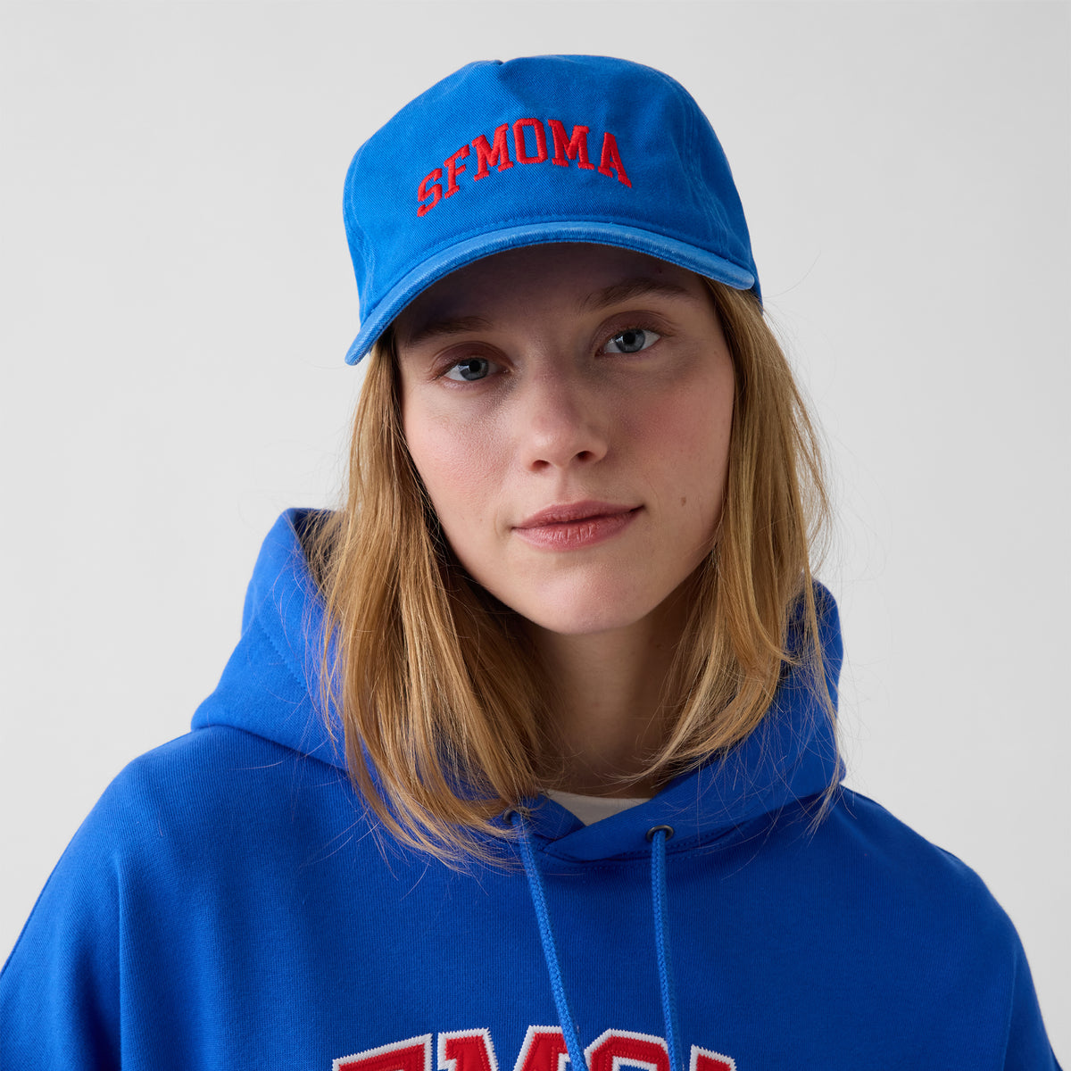 A woman wearing a bright blue baseball cap with red embroidered SFMOMA and a matching blue hoodie, photographed against a plain light gray background.