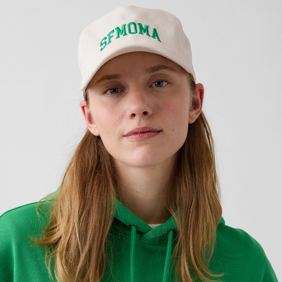 A woman wearing a cream-colored baseball cap with green embroidered 'SFMOMA' text and a matching green hoodie, photographed against a light gray studio background.