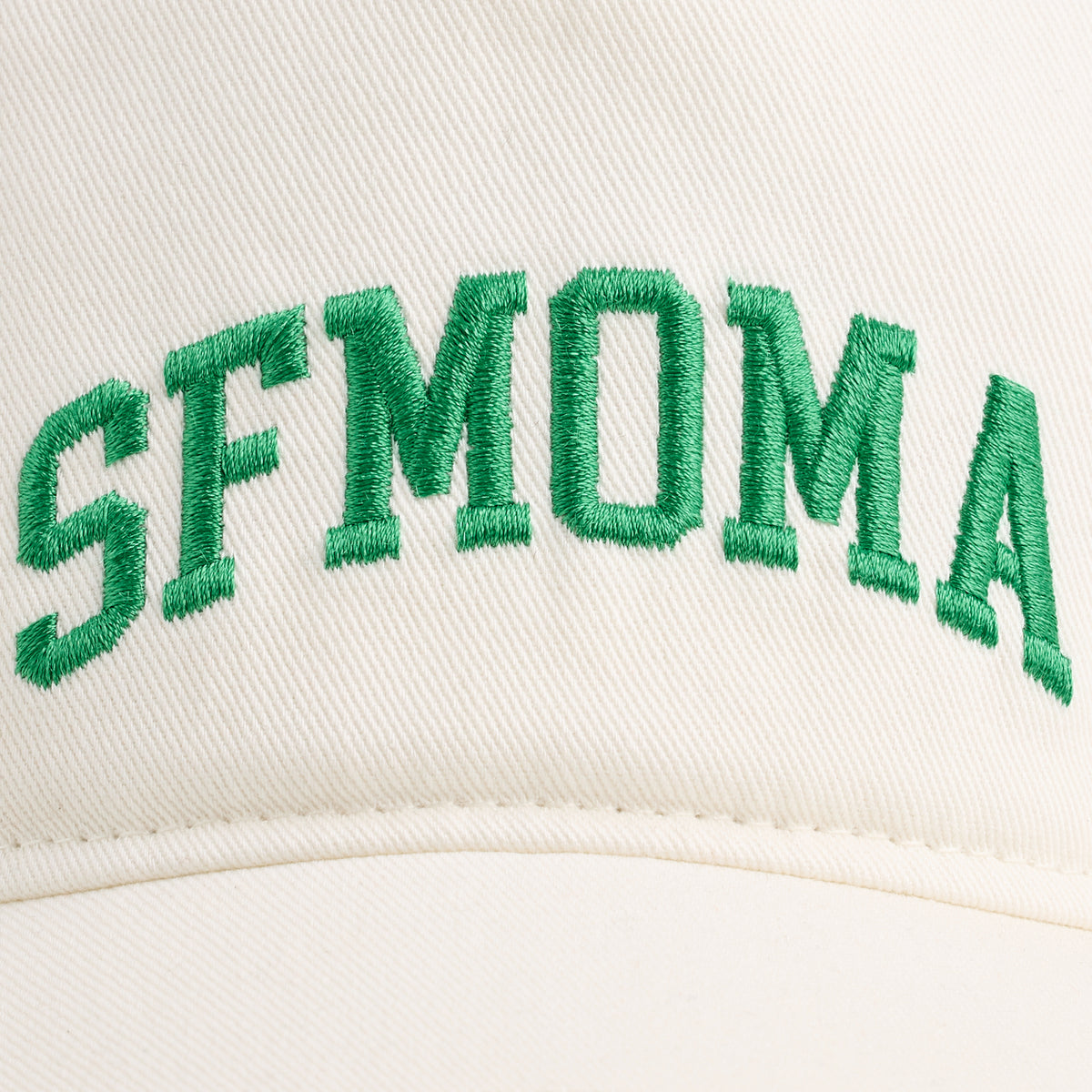 Close-up of a cream-colored baseball cap featuring green embroidered collegiate-style lettering that reads 'SFMOMA' across the front.