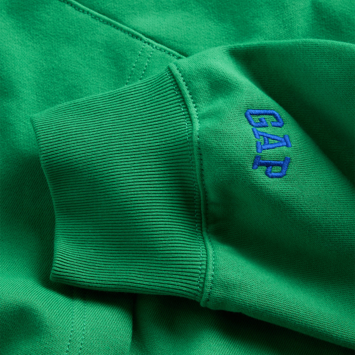Close-up of a green ribbed sweatshirt cuff with a blue embroidered 'GAP' logo on the side.