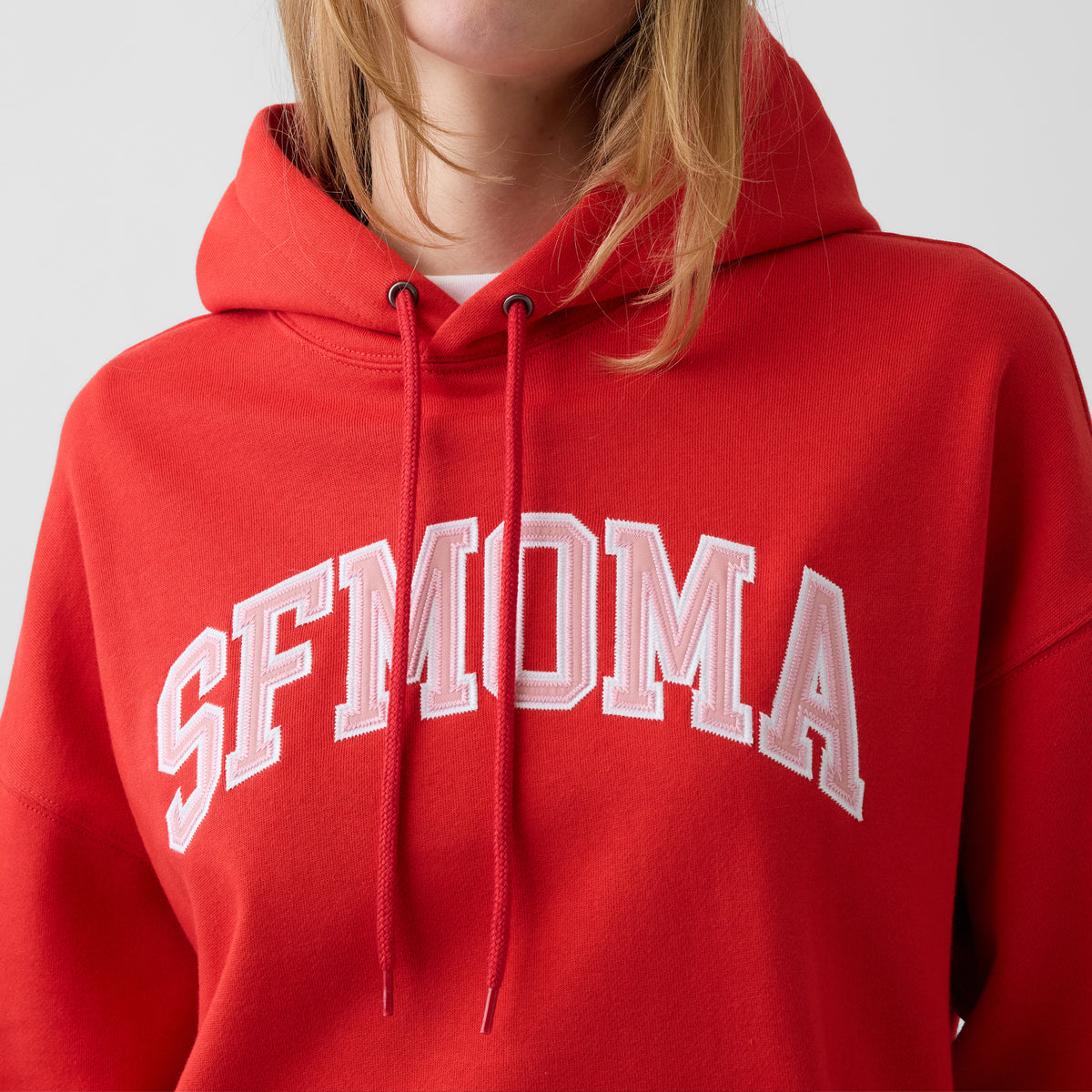A red pullover hoodie with 'SFMOMA' printed in white arched block letters across the chest, featuring a front kangaroo pocket and drawstring hood, shown on a plain white background.