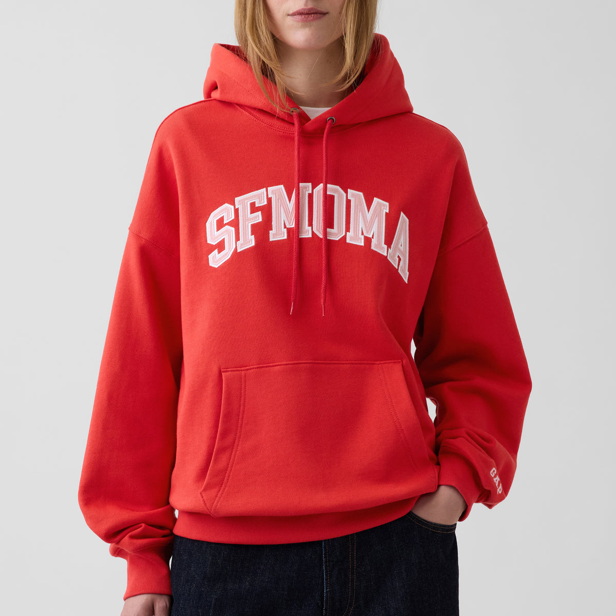 A person wearing a bright red pullover hoodie with 'SFMOMA' printed in white arched letters across the chest, paired with dark jeans against a light gray studio background.
