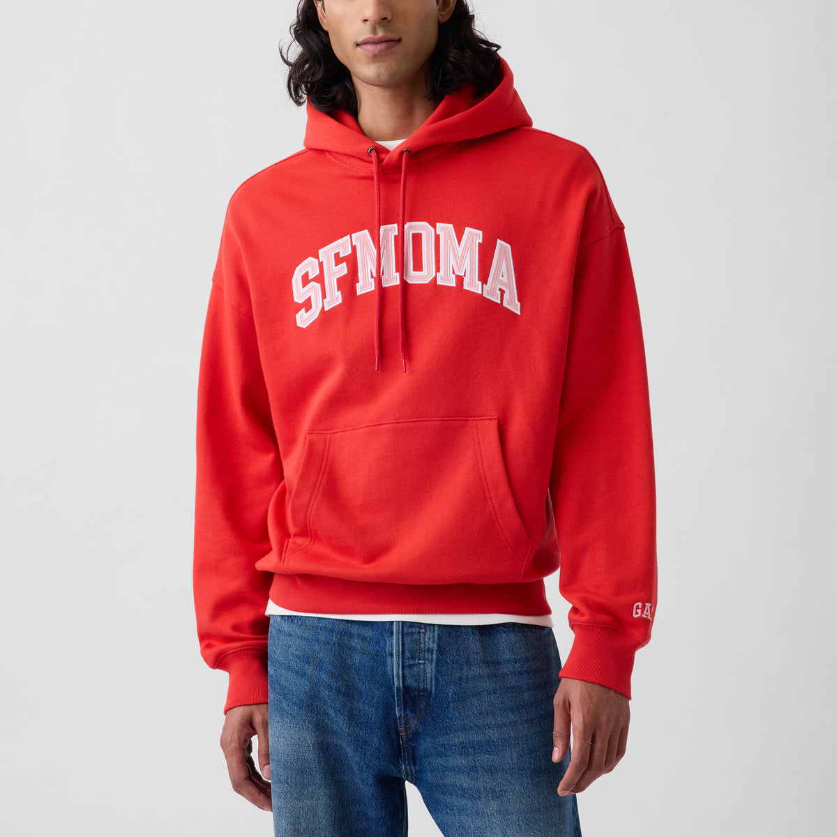 A man wearing a bright red pullover hoodie with 'SFMOMA' printed in white arched letters across the chest, paired with blue jeans, against a light gray studio background.