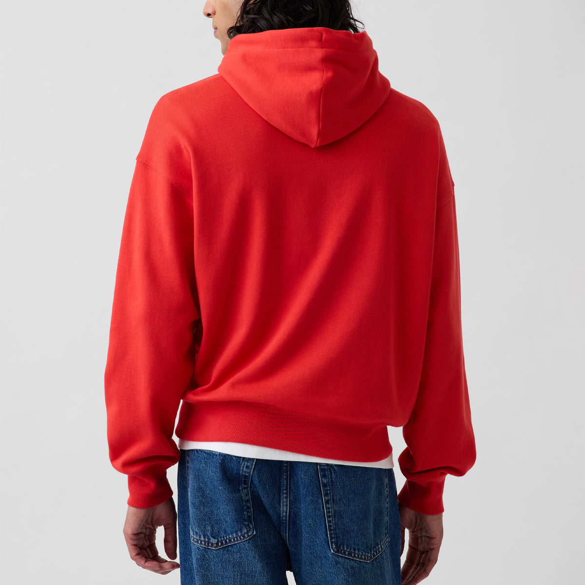 A person shown from the back wearing a bright red pullover hoodie with blue jeans against a plain light gray studio background.
