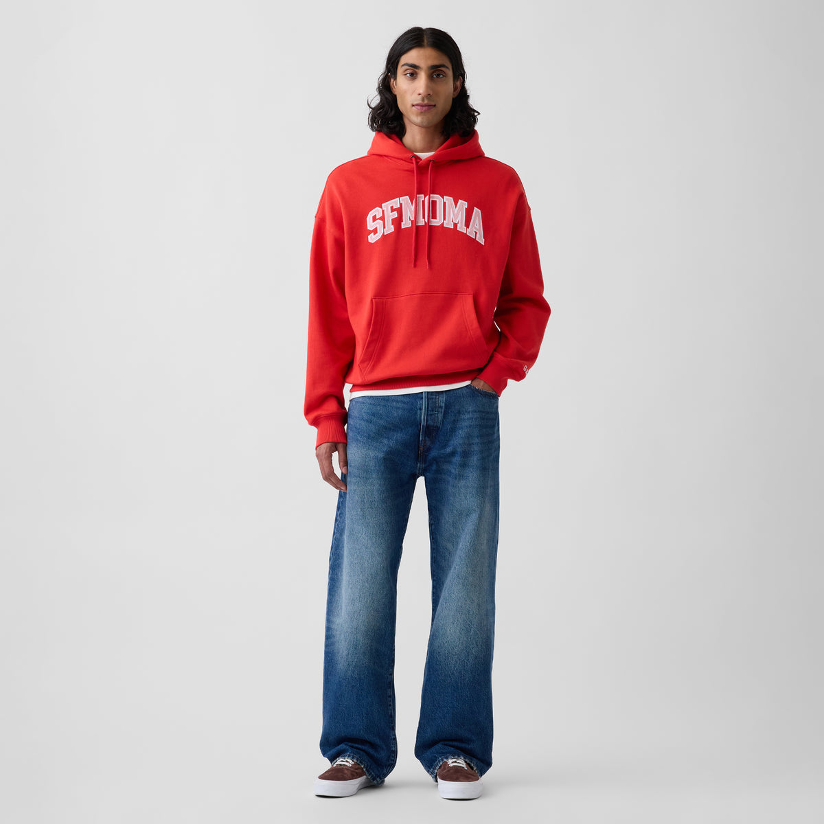 A model stands against a light gray studio background wearing a bright red pullover hoodie with 'SFMOMA' printed in white across the chest, paired with wide-leg blue jeans and brown sneakers.