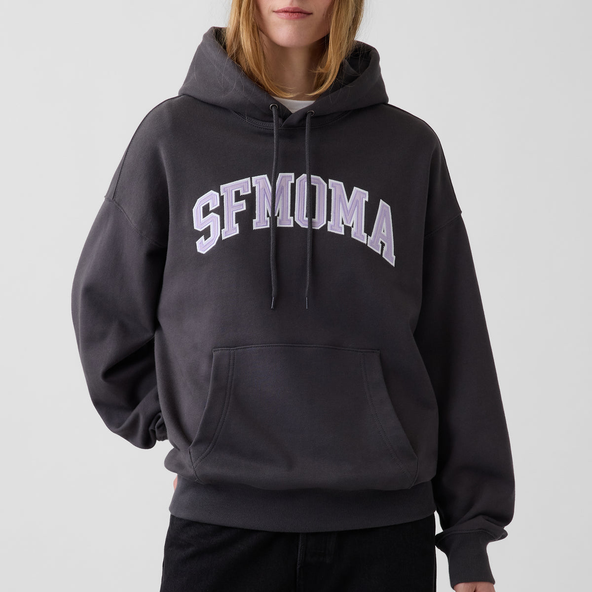 A person wearing a dark charcoal gray pullover hoodie with 'SFMOMA' printed in white arched letters across the chest, shown against a plain light gray studio background.