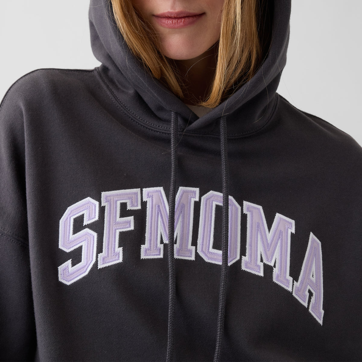 A close-up of a person wearing a dark charcoal gray hoodie with 'SFMOMA' arched across the chest in light lavender and white varsity-style lettering.