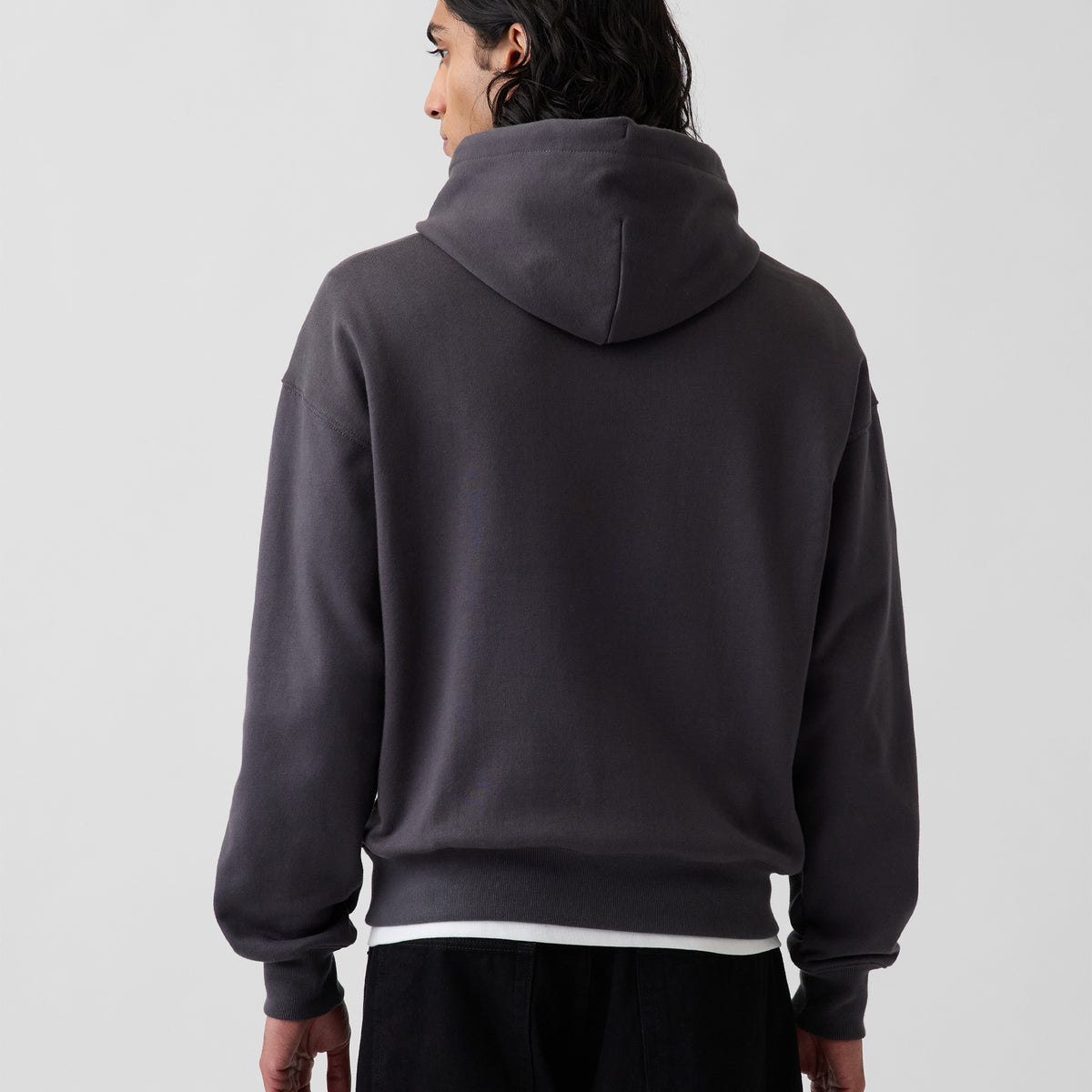 A person shown from the back wearing a dark charcoal gray pullover hoodie with a white undershirt peaking out at the hem, set against a plain light gray studio background.