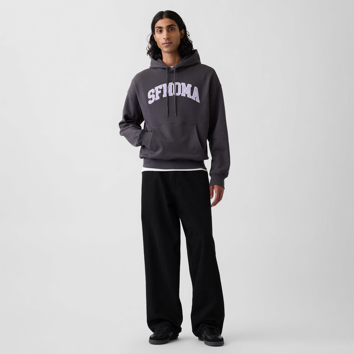 A model stands against a light gray studio background wearing a dark charcoal gray pullover hoodie with 'SFMOMA' printed in white arched text across the chest, paired with black sweatpants and black shoes.