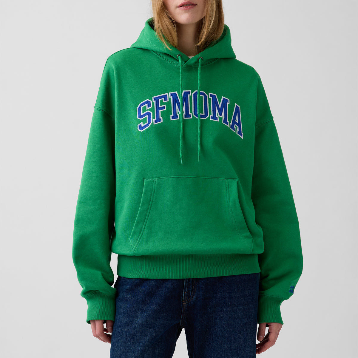 A model wearing a bright green pullover hoodie with 'SFMOMA' printed in blue and white varsity-style lettering across the chest, paired with dark blue jeans against a light gray background.
