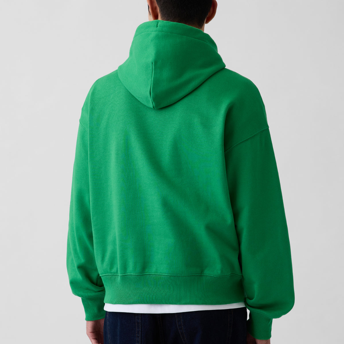 A person viewed from the back wearing a bright green pullover hoodie over a white shirt and dark pants, against a plain light gray studio background.