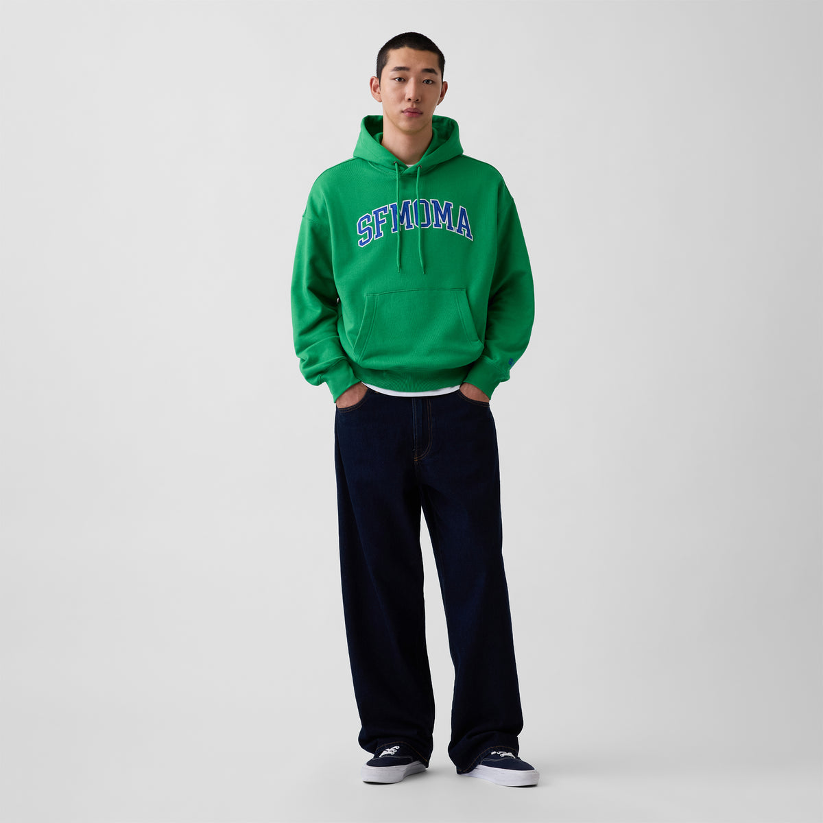 A model stands against a light gray studio background wearing a bright green pullover hoodie with 'SFMOMA' embroidered in blue and white across the chest, paired with dark navy pants and white sneakers.