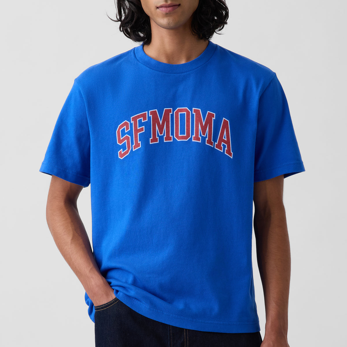 A man wearing a bright blue crewneck t-shirt with 'SFMOMA' printed in red varsity-style letters with a white outline across the chest, set against a plain light gray background.