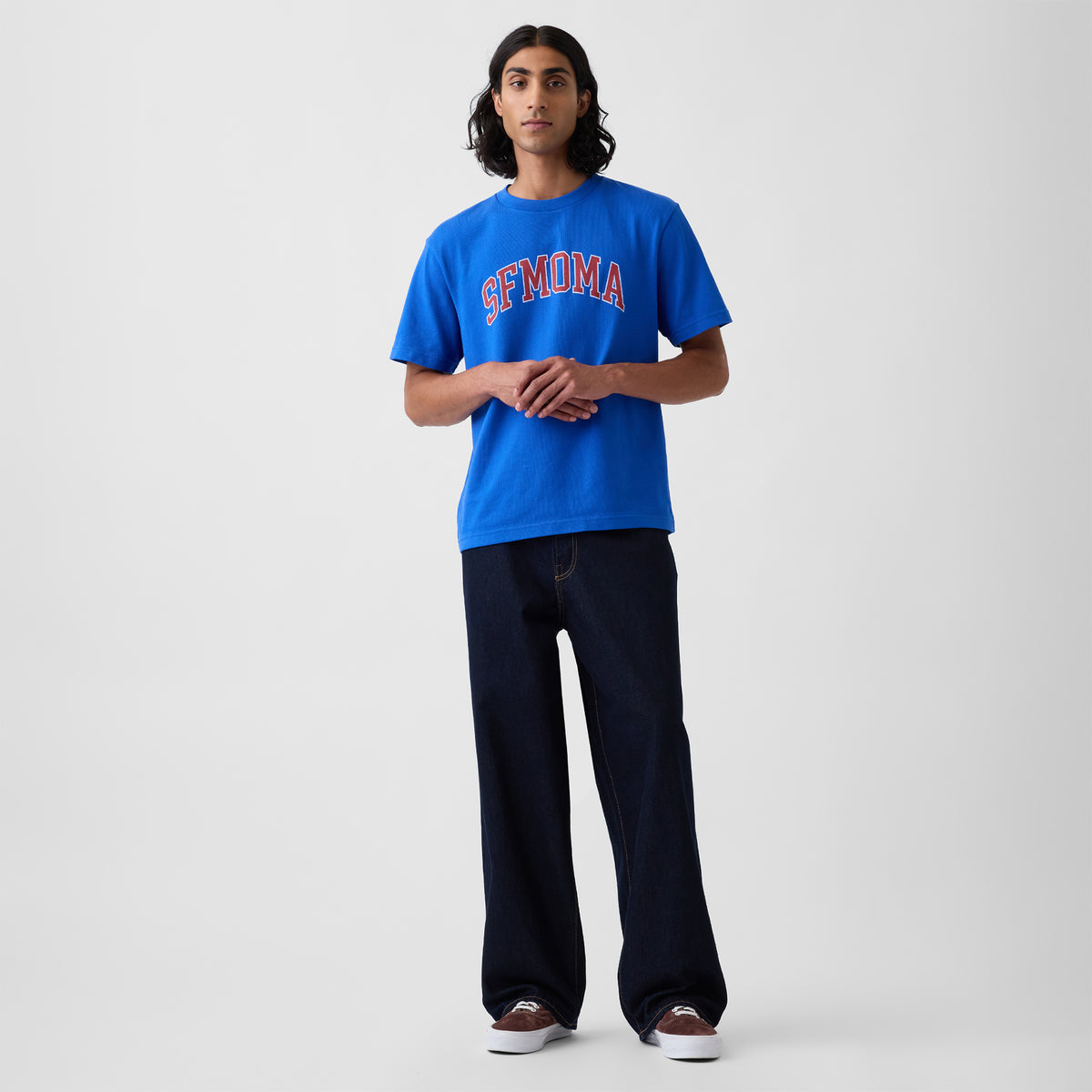A model stands against a light gray studio background wearing a bright blue crewneck t-shirt with 'SFMOMA' printed in red and white across the chest, paired with dark wide-leg jeans and brown sneakers.
