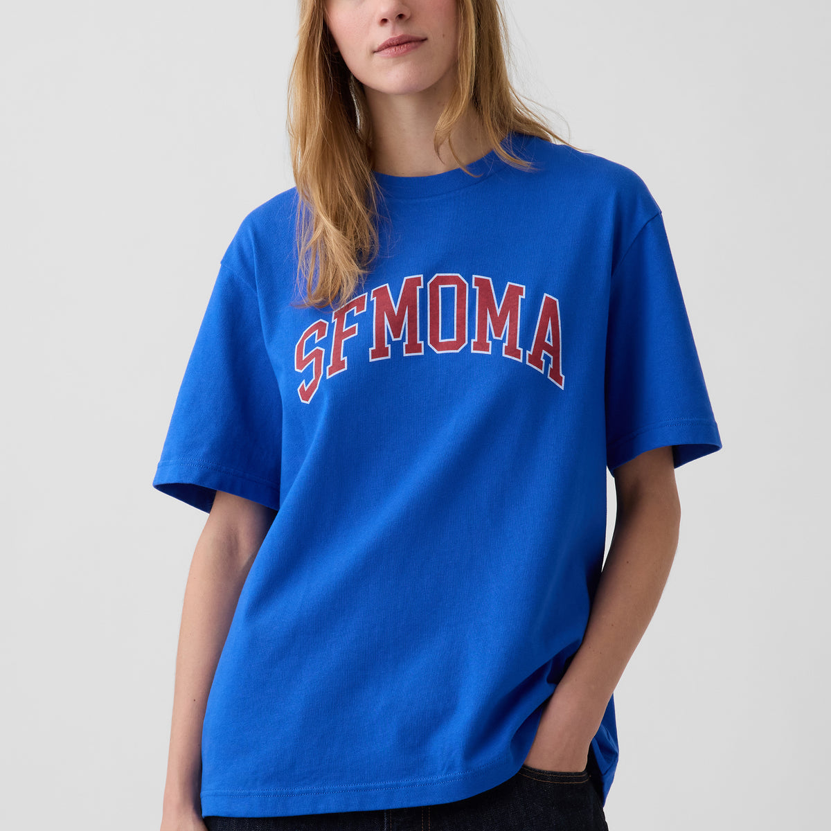 A woman wearing a bright blue short-sleeve t-shirt with 'SFMOMA' printed in red arched letters with a white outline across the chest, set against a plain light gray background.