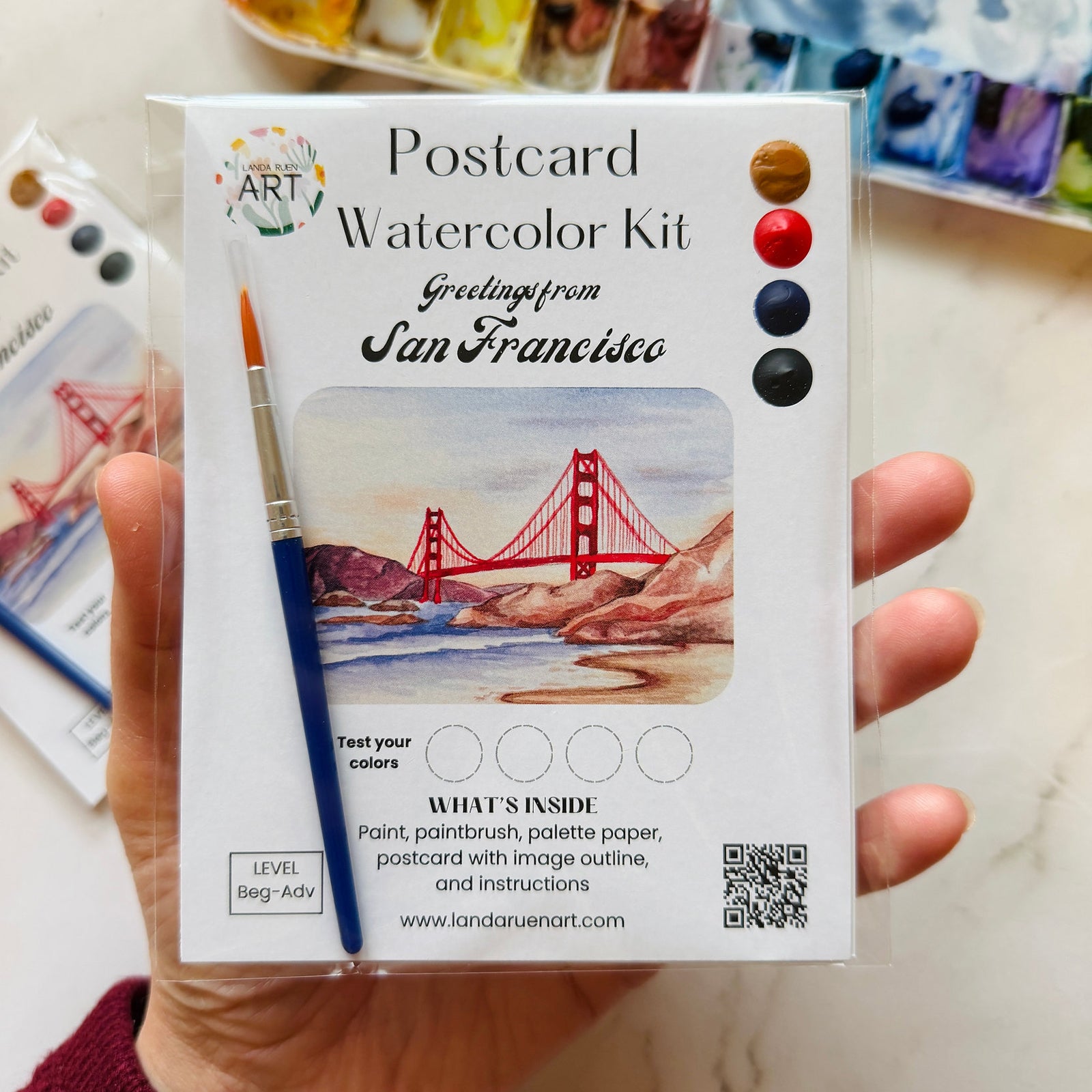 A hand holding a packaged watercolor postcard kit labeled 'Postcard Watercolor Kit' with 'Greeting from San Francisco' and an illustration of the Golden Gate Bridge. The package includes paint color swatches, a paintbrush, and product details on a light marble-like surface.