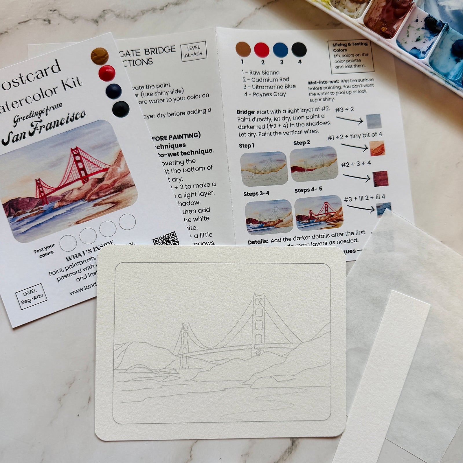 A hand holding a packaged watercolor postcard kit labeled 'Postcard Watercolor Kit' with 'Greeting from San Francisco' and an illustration of the Golden Gate Bridge. The package includes paint color swatches, a paintbrush, and product details on a light marble-like surface.