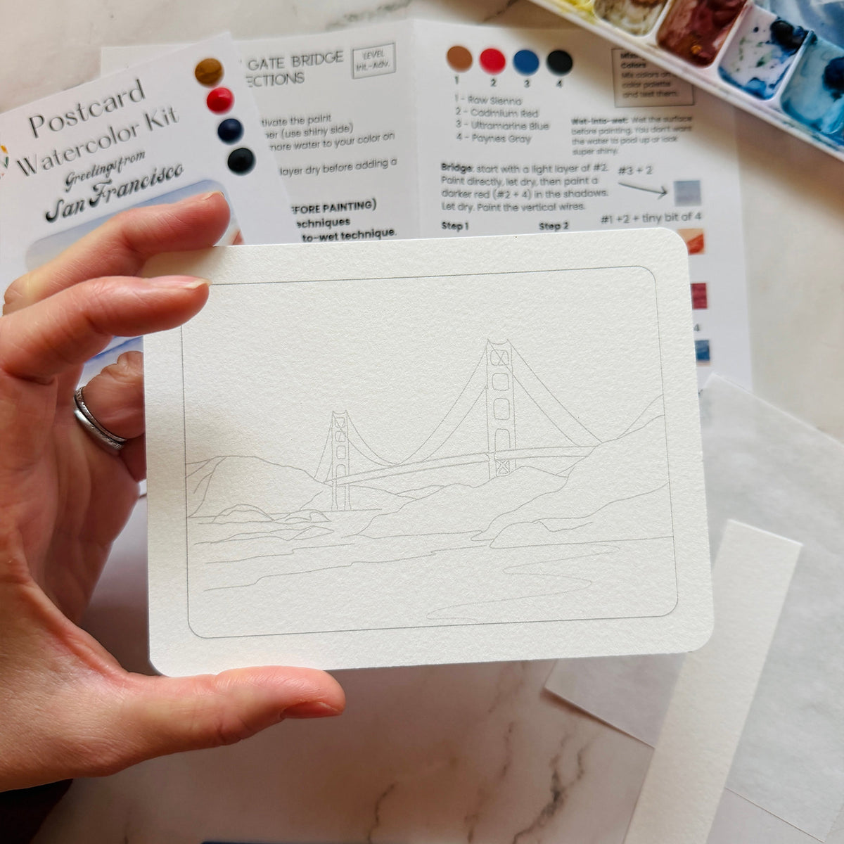 A hand holds a white postcard featuring a black ink line drawing of the Golden Gate Bridge. In the background, printed watercolor painting instructions and paint swatches are visible on a white surface.