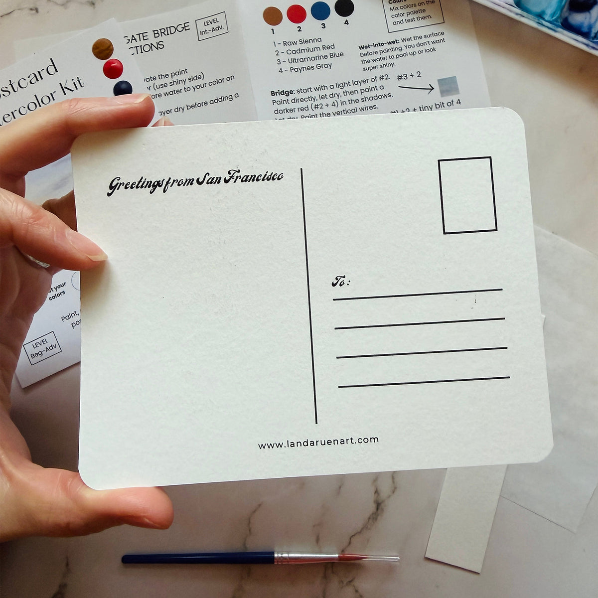 A hand holds a white postcard printed with 'Greetings from San Francisco' and postcard lines, with a Landa Ruen Art color kit sheet and paintbrush visible in the background on a marble surface.