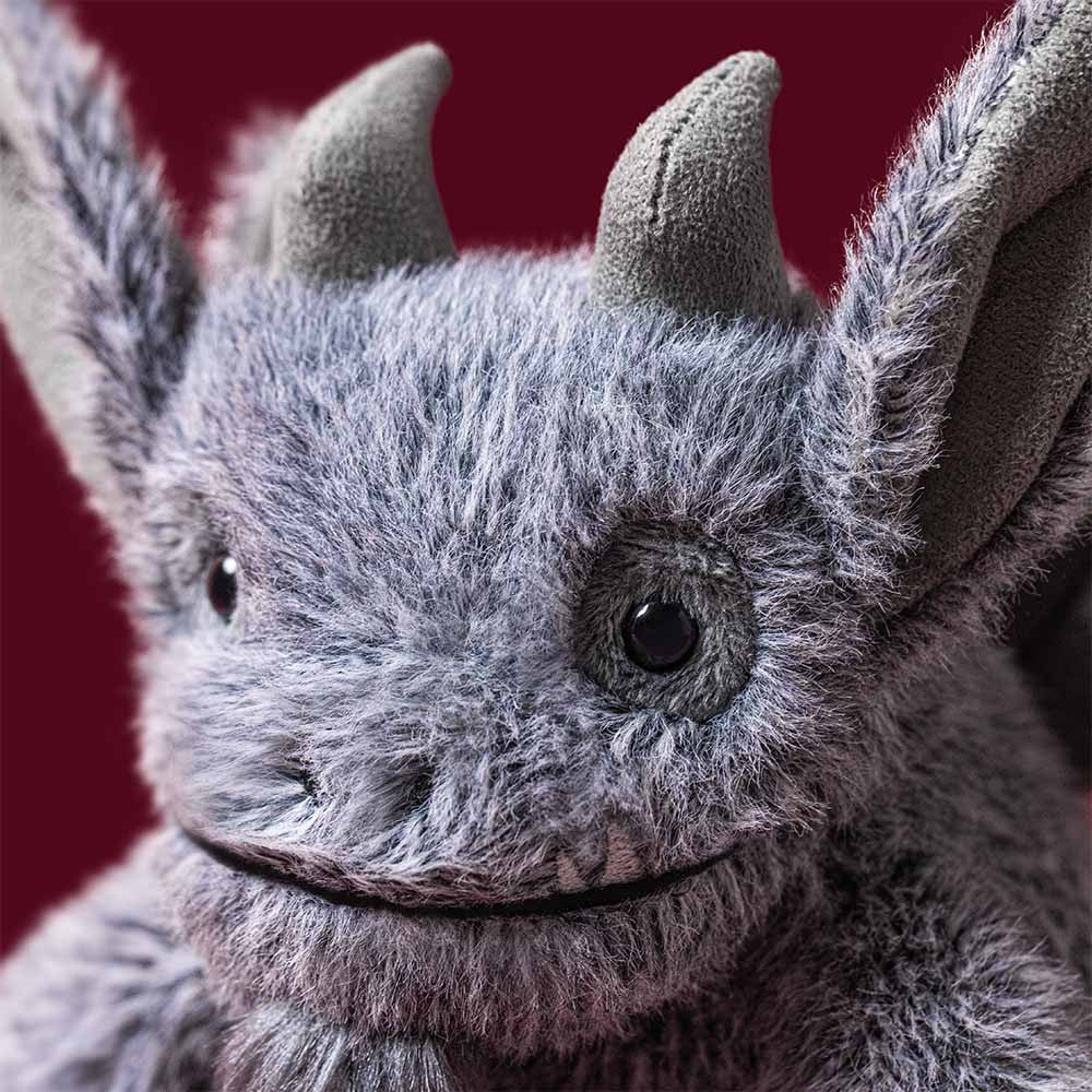 A close-up of a gray furry monster toy with pointed ears, dark eyes, and a stitched smile against a dark red background.
