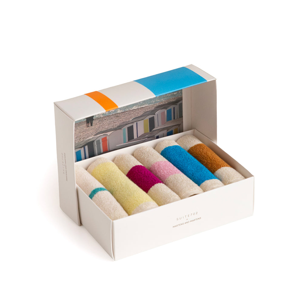 An open white gift box containing six rolled bath hand towels in assorted pastel and bright colors, with a printed beach scene on the inside lid. 