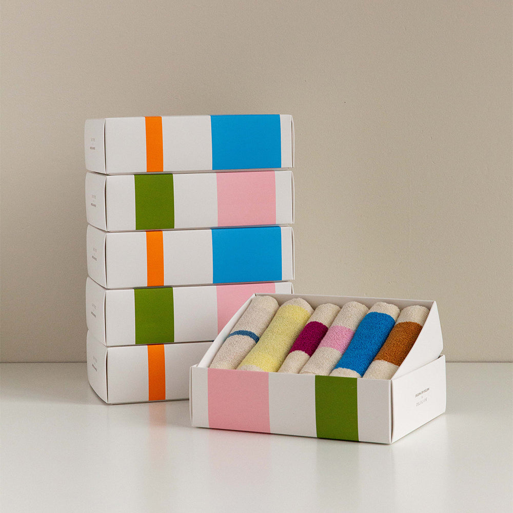 A set of white gift boxes with colorful striped and solid color panels, with one open box displaying multiple rolled cream-colored socks or fabric rolls in pastel and bright colors, on a white surface against a beige background.