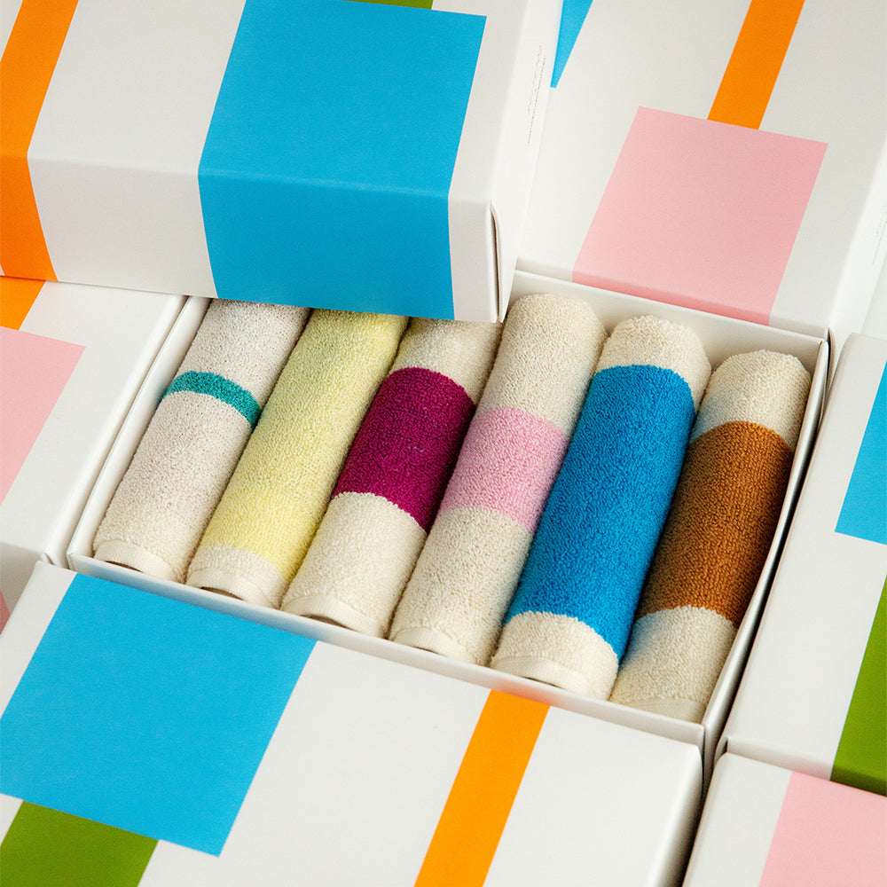 A set of six rolled hand towels in cream, yellow, magenta, pink, blue, and brown, arranged inside an open white gift box with colorful geometric patterned wrapping paper.