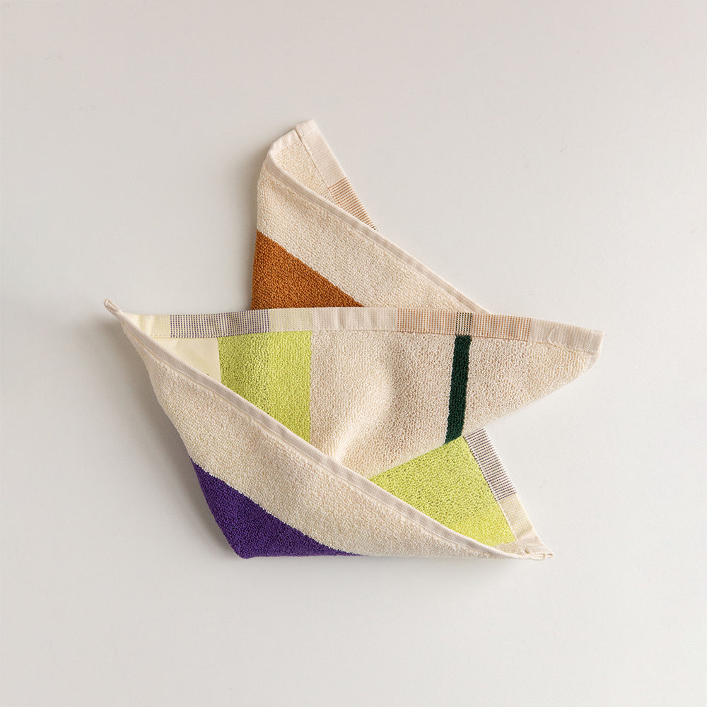 A folded cotton hand towel with a geometric color-block pattern in cream, lime green, purple, orange, and dark green, displayed on a light gray background.