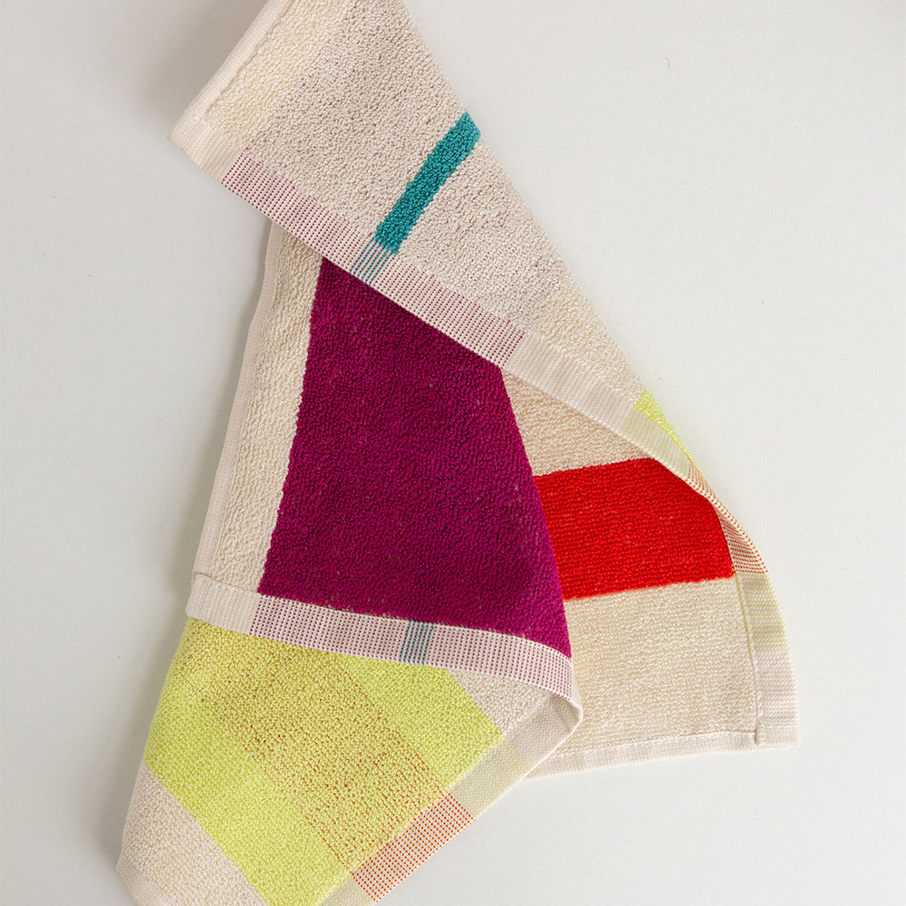 A folded cotton towel with a colorful geometric pattern of magenta, red, yellow, and teal blocks on a cream background, photographed on a plain white surface.