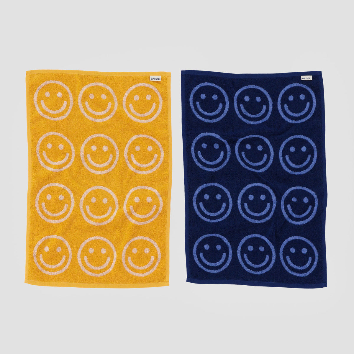 Two towels front side, one beige with yellow smiley faces and one blue with navy smiley faces, on a light gray background.