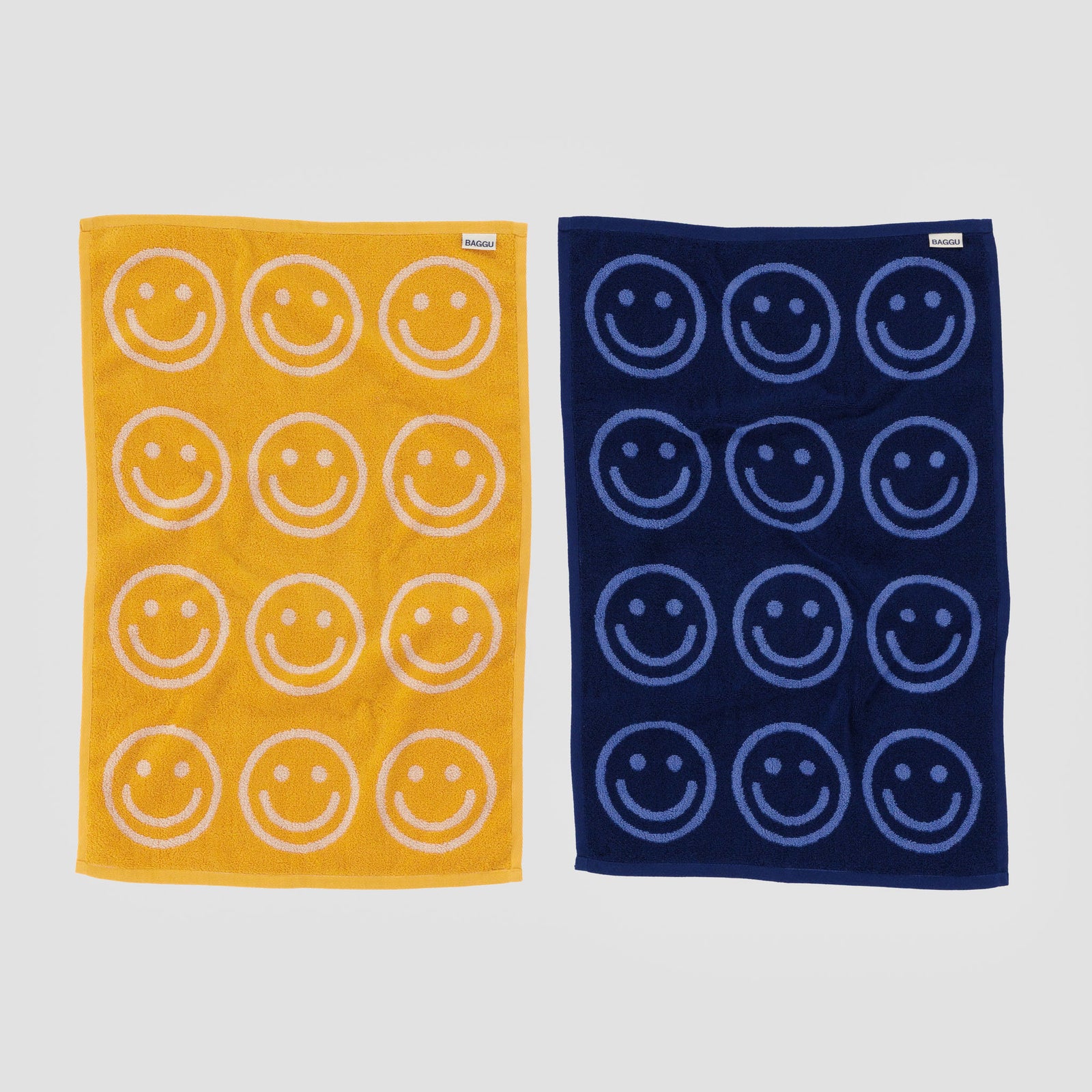 Two folded towels, one blue with navy patterns and one yellow with marigold patterns, on a white background. Both towels have 'Baggu' branding.
