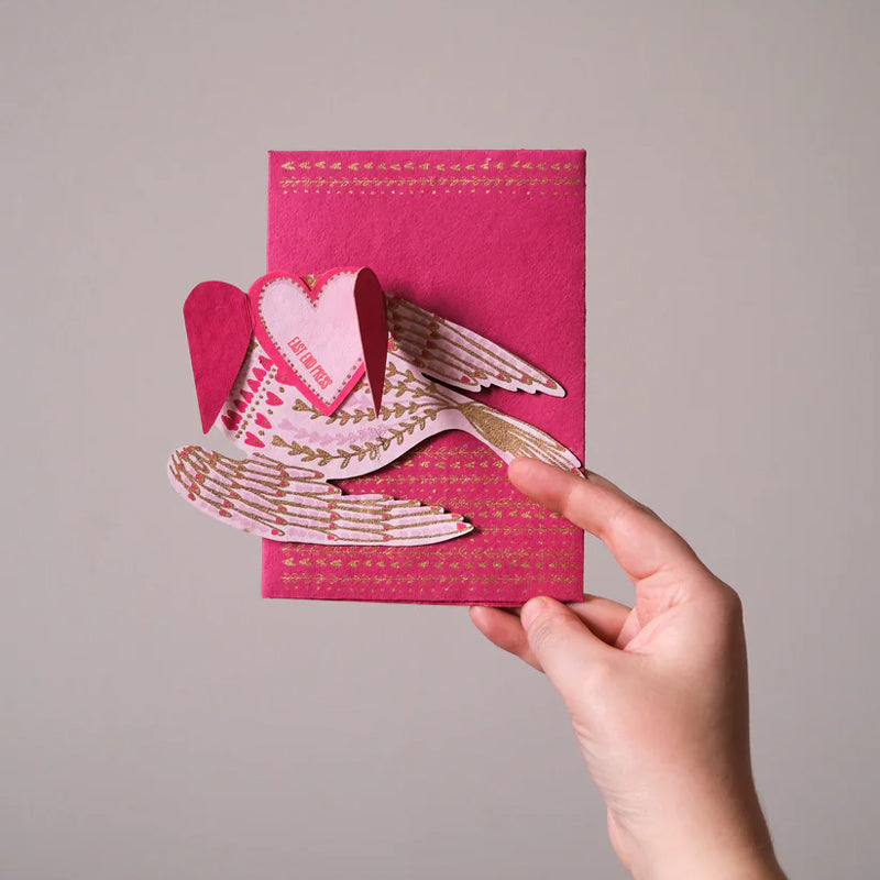 Hand holding a pink card with a bird and heart design against a gray background