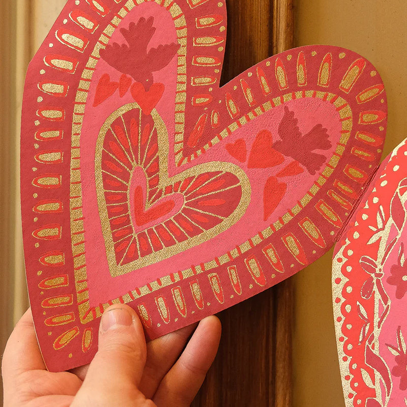 Hand holding a decorative pink heart card with gold and red patterns.