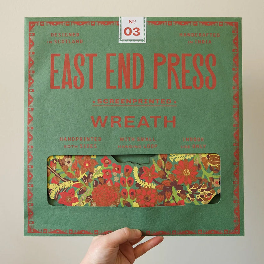 A hand holds a green square envelope labeled 'EAST END PRESS WREATH' with a floral-patterned window revealing a red and yellow wreath inside, against a plain light wall.