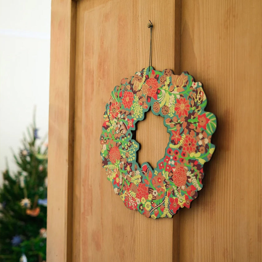 A colorful floral wooden wreath hanging on a light brown wooden door, with a blurred Christmas tree in the background.