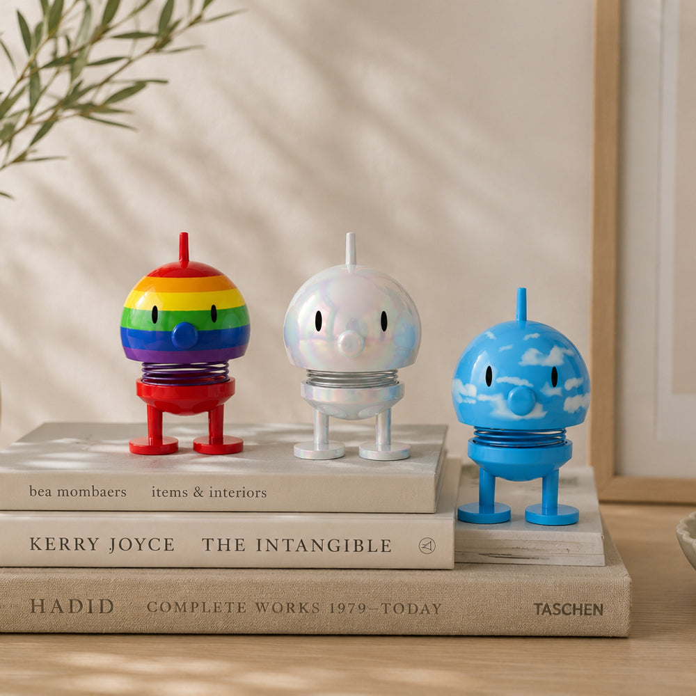 Three small decorative toy figures in rainbow, iridescent white, and blue sky designs, displayed on stacked books in a styled indoor setting.
