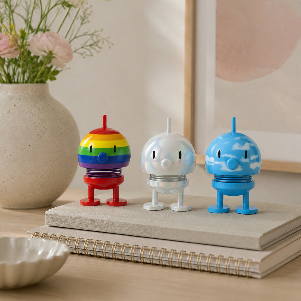 Three small decorative toy figures in rainbow, iridescent white, and blue sky designs, displayed on stacked books in a styled indoor setting.