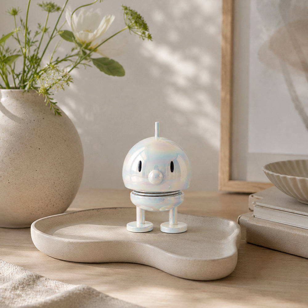 An iridescent white figurine with a metallic, holographic finish, featuring a round head, simple black eyes, a small protruding antenna, a spring-like neck, and two round legs, centered on a solid white background.