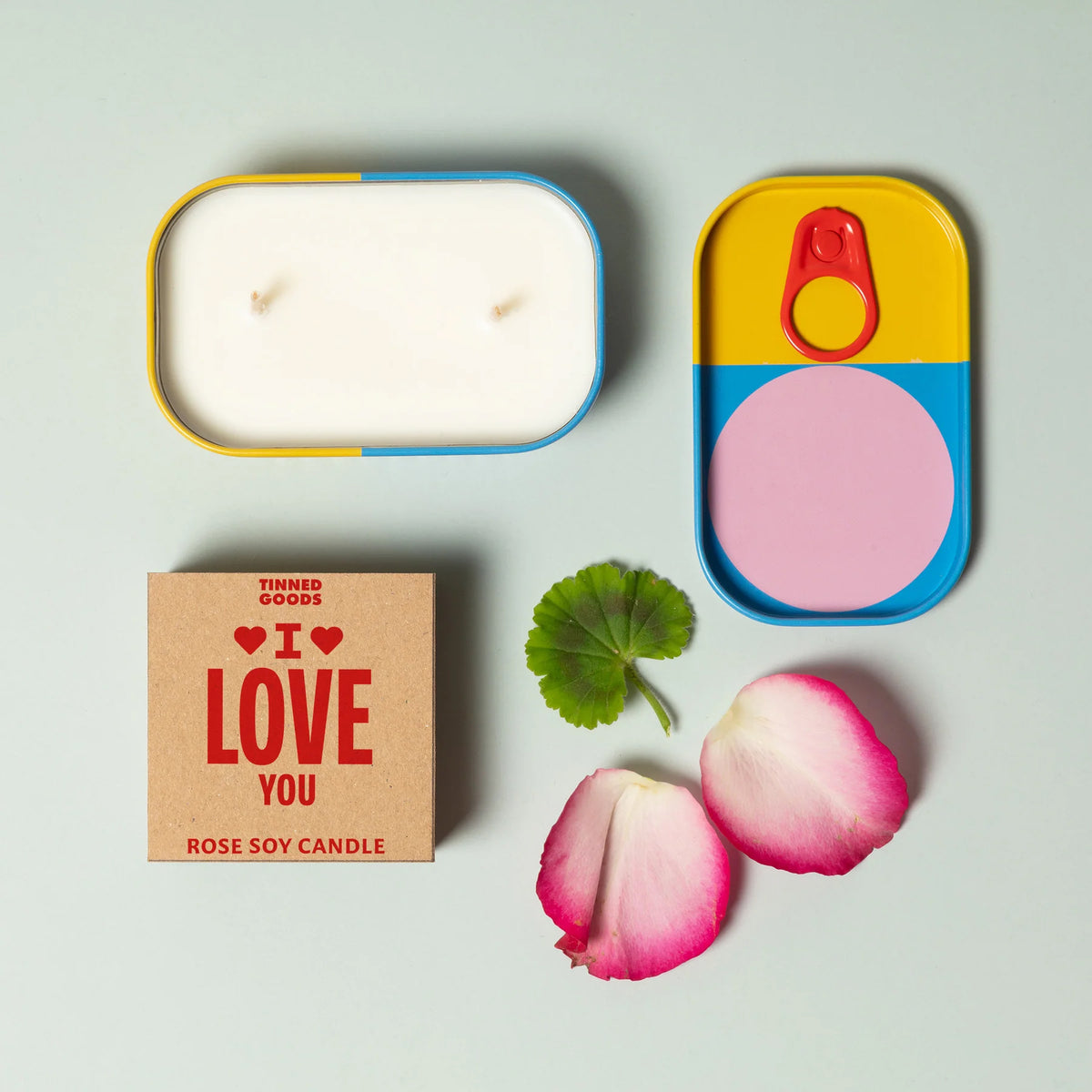 Candle in a tin with decorative lid, 'I LOVE YOU' box, and rose petals on a light gray background