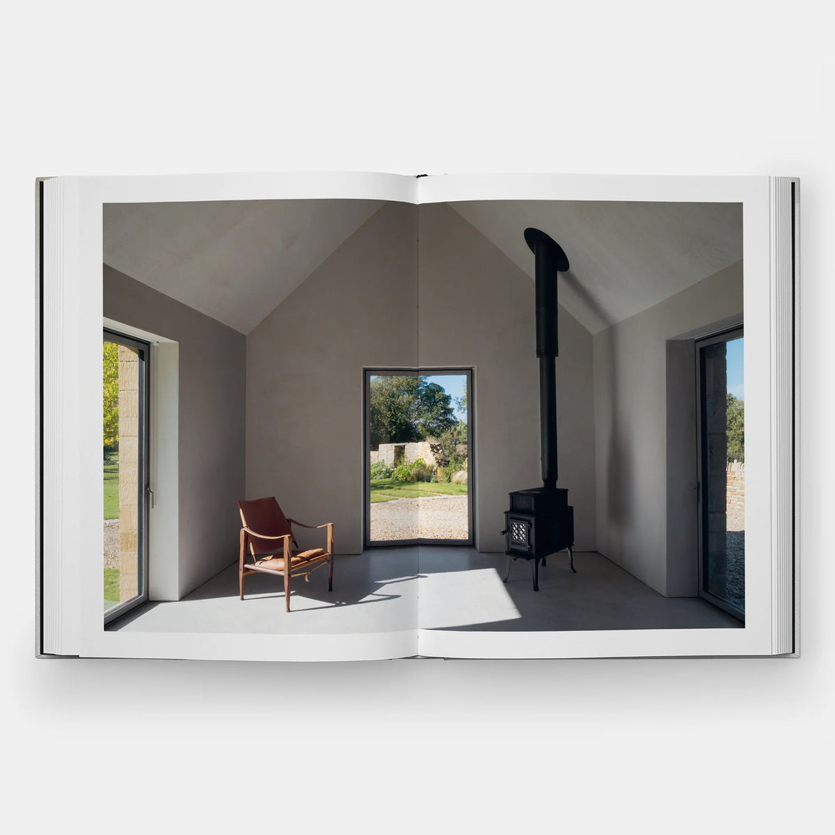 An open book displaying a high-quality interior architectural photograph of a minimalist room with a slanted ceiling, a brown leather chair, and a black wood stove, set against a clean white background.