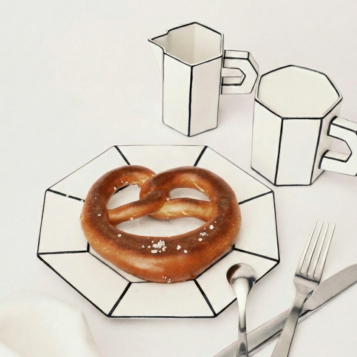 Pretzel on a geometric plate with geometric coffee pots and cutlery on a white background