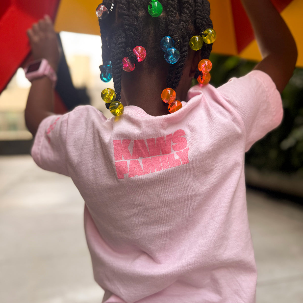 Child wearing a pink 'Kaws Family' t-shirt with colorful beads in hair, blurred background