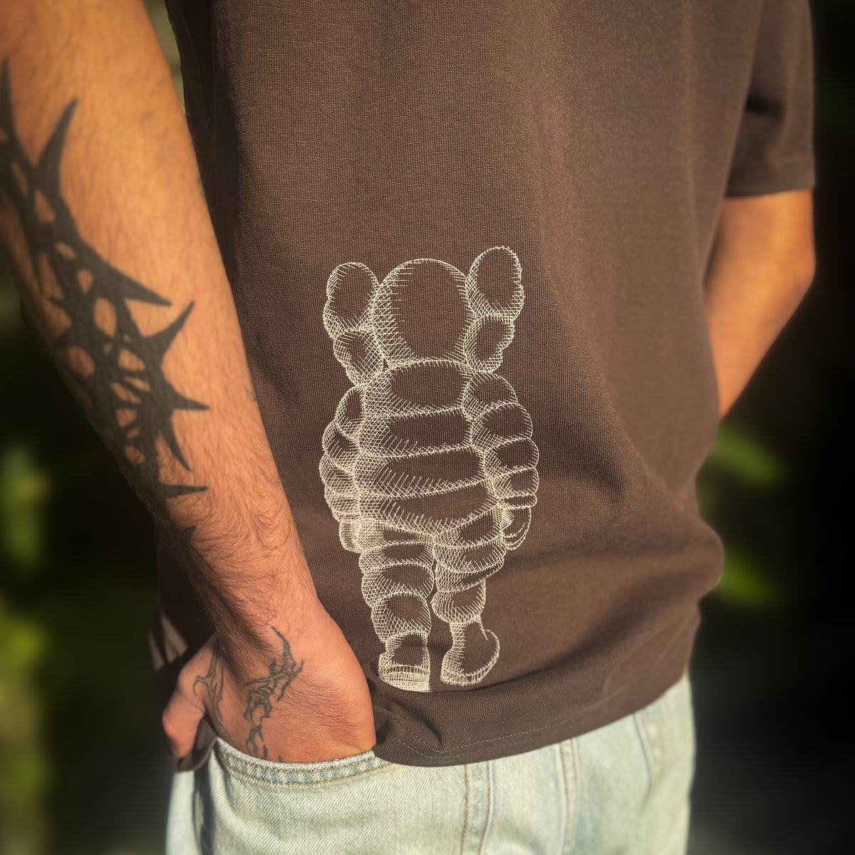 Person wearing a brown t-shirt with a white KAWS design, standing outdoors.